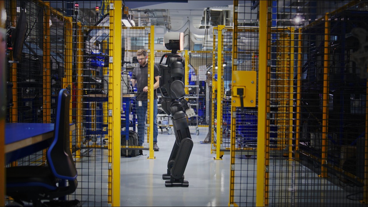 Boston Dynamics’ Atlas standing in a caged prototype shop, with a technician behind it.