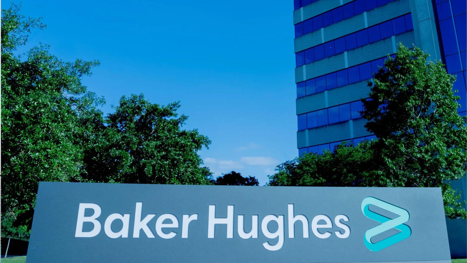 The Baker Hughes name and logo on a sign in front of trees and a glass office tower.