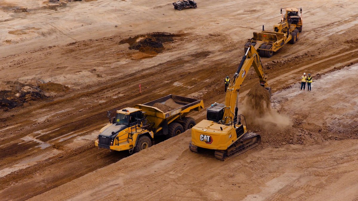 A yellow excavator loading soil into a truck on a wide construction site.
