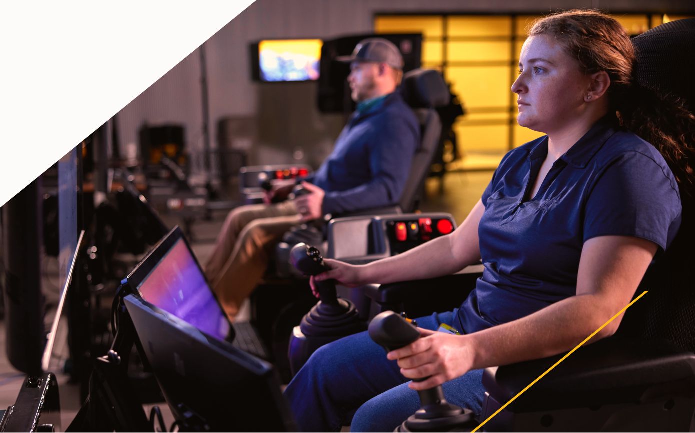 Two people in a dark training room sit in equipment simulators, each holding a control stick, with screens in front of them.