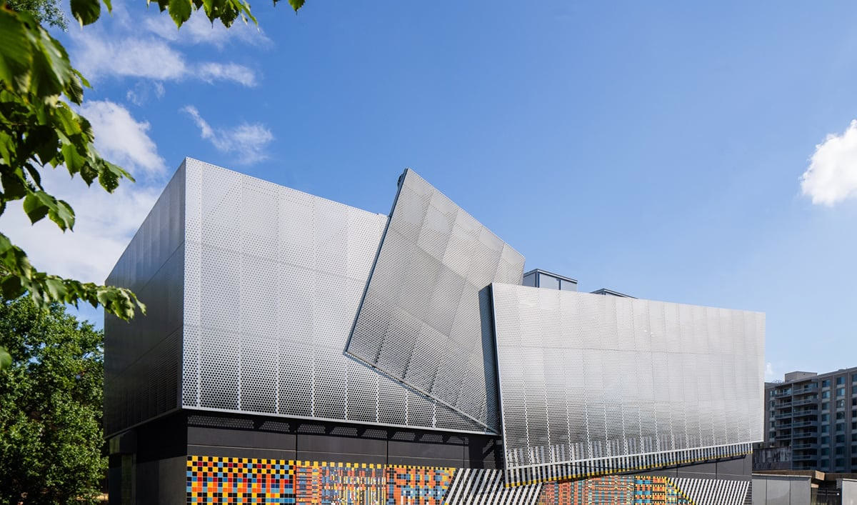 A silver metal building with a perforated facade and a band of coloured ceramic tiles along the base, under a blue sky.