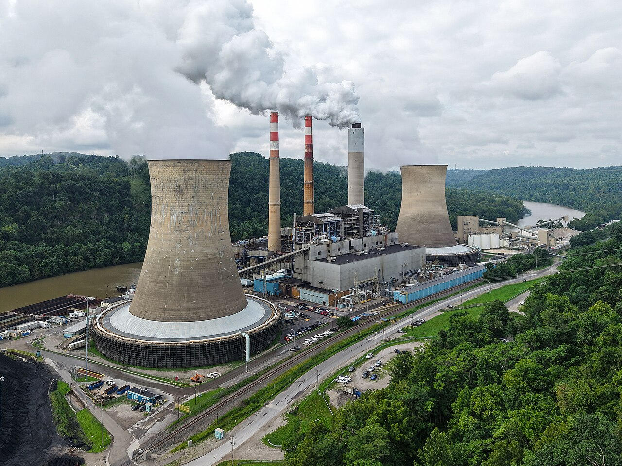 An aerial view of a coal-fired power station with two large cooling towers, striped chimneys and a river, set against forested hills.