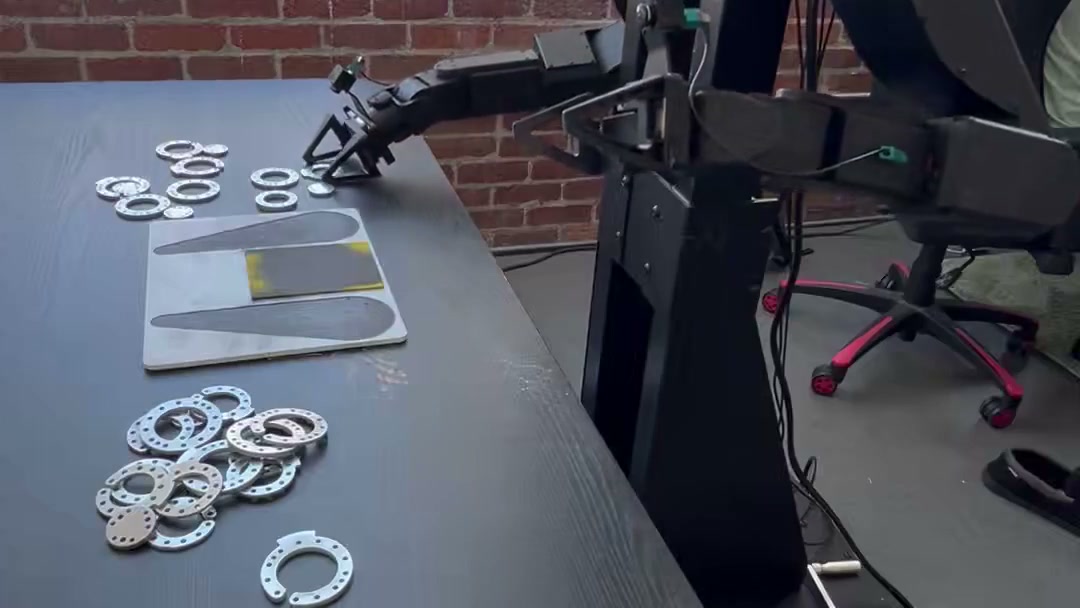 A robot arm from Physical Intelligence’s API post, working over a table of metal parts.