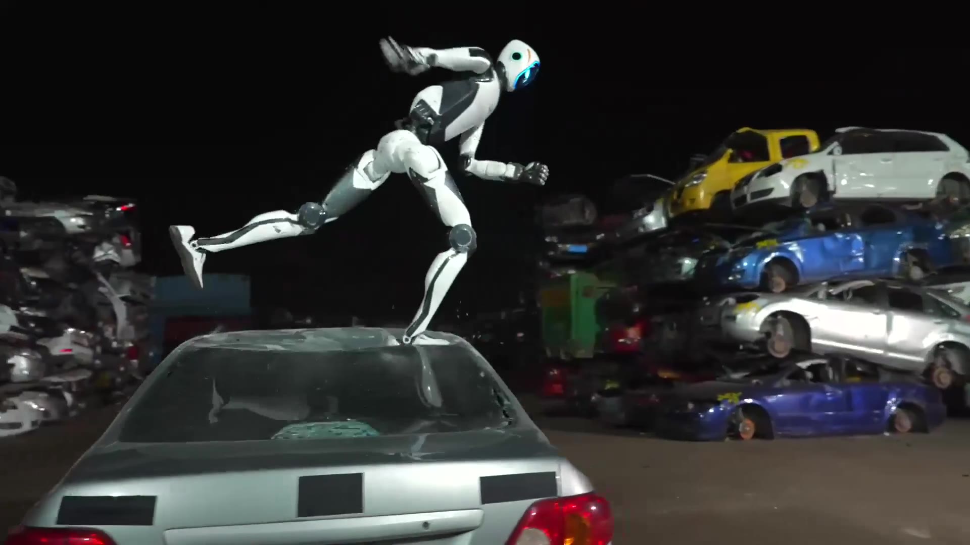 A white humanoid robot mid-jump above the roof of a car in a scrapyard at night.